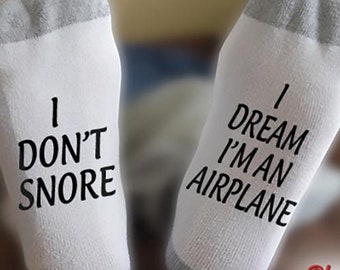 May include: A pair of white socks with black text. One sock says "I don't snore" and the other says "I dream I'm an airplane".