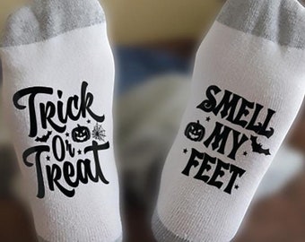 May include: A pair of white socks with black text. One sock says "Trick or Treat" with a pumpkin and spider web graphic. The other sock says "Smell My Feet" with a pumpkin and bat graphic.