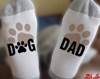 May include: White socks with gray heels and toes. One sock has a tan paw print with the word "DOG" in black. The other sock has a tan paw print with the word "DAD" in black. The socks are a fun gift for dog lovers.