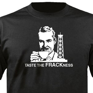 May include: Black t-shirt with a white graphic of a man drinking from a cup, a drilling rig, and the text "TASTE THE FRACKNESS". The shirt is made of a soft material and has short sleeves.