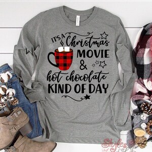 May include: Gray long-sleeve shirt with the text "It's a Christmas Movie & hot chocolate kind of day" in black. A red and black plaid mug with marshmallows is pictured next to the text.
