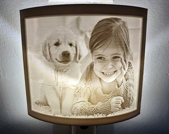 May include: A white ceramic night light with a 3D image of a girl and a golden retriever puppy. The image is in a rectangular frame and the night light has a plug-in cord.
