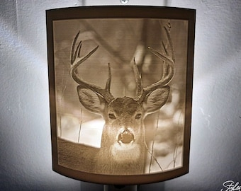 May include: A white ceramic night light with a 3D image of a white-tailed deer with antlers. The deer is looking directly at the camera. The night light is illuminated and the deer is visible in the light.