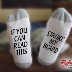 May include: A pair of white socks with gray heels and toes. One sock reads "IF YOU CAN READ THIS" in black text, and the other reads "STROKE MY BEARD". The socks are a novelty item.