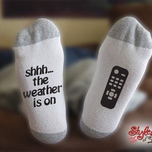 May include: A pair of white socks with a grey cuff. One sock has the text "shhh... the weather is on" printed on it. The other sock has a black and white image of a remote control printed on it.