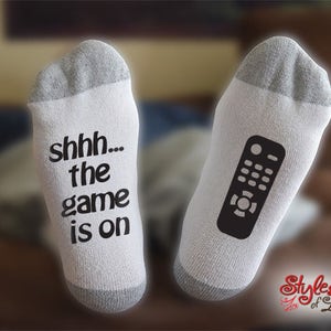 May include: A pair of white socks with a grey trim. One sock has the text "shhh... the game is on" printed on it. The other sock has a black and white image of a remote control printed on it.