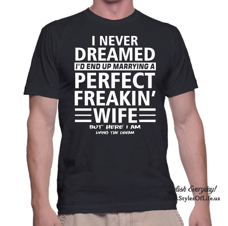 May include: Black t-shirt with white text. The text reads: "I NEVER DREAMED I'D END UP MARRYING A PERFECT FREAKIN' WIFE BUT HERE I AM LIVING THE DREAM." The shirt has a crew neck and short sleeves. The text is in a bold, sans-serif font.
