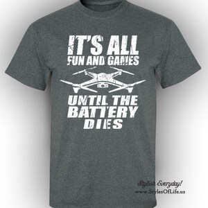 May include: A dark grey t-shirt with white text that reads "It's all fun and games until the battery dies." A white silhouette of a drone is above the text. The text "Stylish Everyday!" and a website address are at the bottom of the shirt.