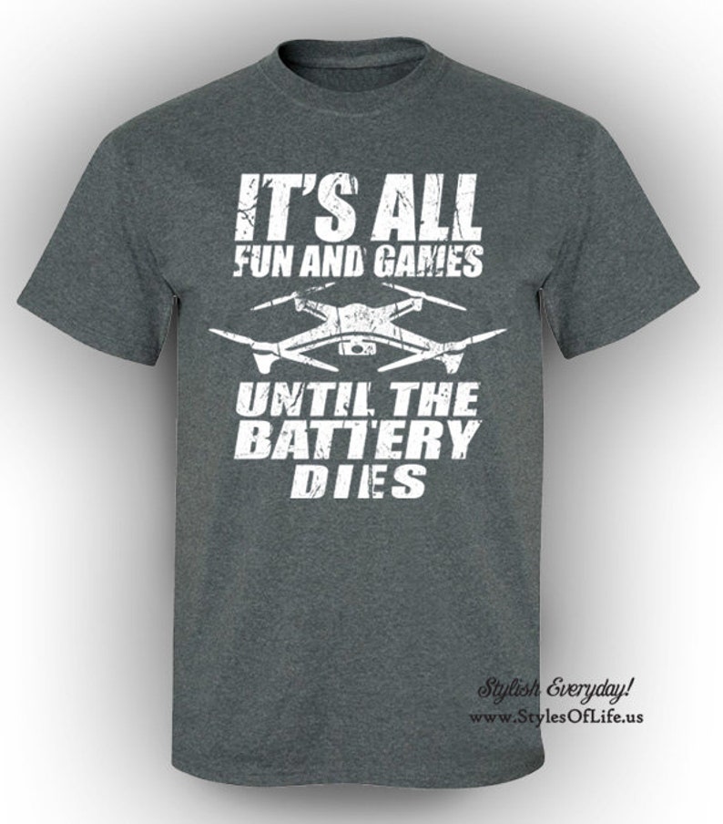 May include: A dark grey t-shirt with white text that reads "It's all fun and games until the battery dies." A white silhouette of a drone is above the text. The text "Stylish Everyday!" and a website address are at the bottom of the shirt.