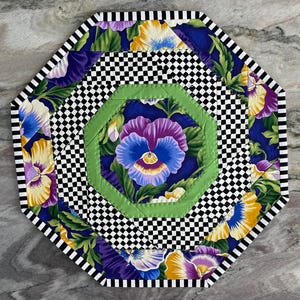 Può includere: Un sottopentola trapuntato ottagonale con un motivo floreale di viole del pensiero nei toni del viola, giallo e blu. Il sottopentola ha un motivo a scacchi bianchi e neri e un bordo verde, con un bordo a righe. Lo sfondo è blu scuro.