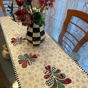 Puede incluir: Un camino de mesa blanco con un borde a cuadros blanco y negro. El camino de mesa está decorado con un patrón de copos de nieve y tres árboles de Navidad aplicados en tonos verdes, rojos y negros.