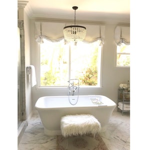 May include: A white bathtub with a chrome faucet and a white fluffy ottoman in a bathroom with a white marble floor and a chandelier with crystal beads.