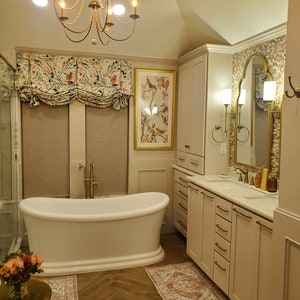 May include: A white freestanding bathtub with gold fixtures in a bathroom with white cabinets, a white countertop, and a gold mirror. The bathroom has a beige tiled floor and a patterned rug.