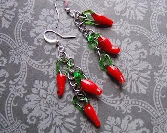 May include: A pair of dangling earrings featuring red chili pepper charms. Each earring has a silver-tone hook and chain, with multiple red peppers and green accents. The earrings are set against a gray floral patterned background.