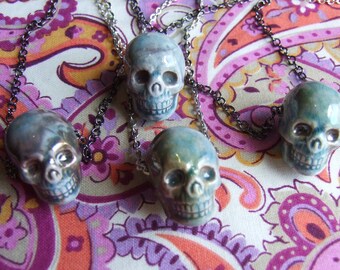 May include: Four ceramic skull pendants on silver chains, hanging on a paisley patterned fabric background. The skulls are painted in shades of blue and green.