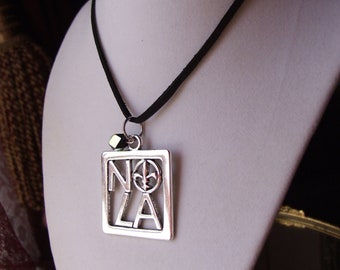 Puede incluir: Un collar de plata con un cordón negro y un colgante que dice "NOLA" con un diseño de flor de lis. El colgante está en un marco cuadrado.
