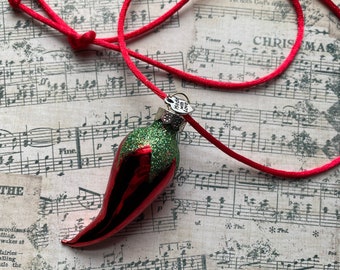 May include: Red glass Christmas ornament shaped like a chili pepper with green glitter and a silver charm that says "Made with Love". The ornament is hanging from a red cord on a sheet of music.