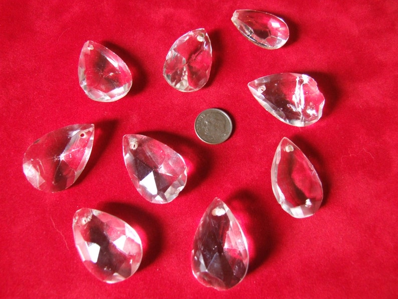 Puede incluir: Diez prismas de cristal transparentes en forma de l&aacute;grima con un dise&ntilde;o facetado, presentados sobre una superficie de terciopelo rojo.