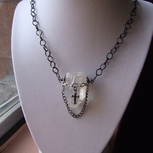 May include: A silver chain necklace with a clear quartz crystal pendant. The pendant is surrounded by a smaller silver chain and features a small silver cross.