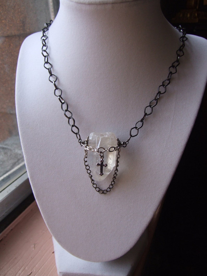 May include: A silver chain necklace with a clear quartz crystal pendant. The pendant is surrounded by a smaller silver chain and features a small silver cross.
