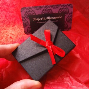 May include: A small black gift box with a red ribbon tied in a bow. The box is sitting on a red tissue paper background. The box has the text "Majestic Menagerie" printed on it.
