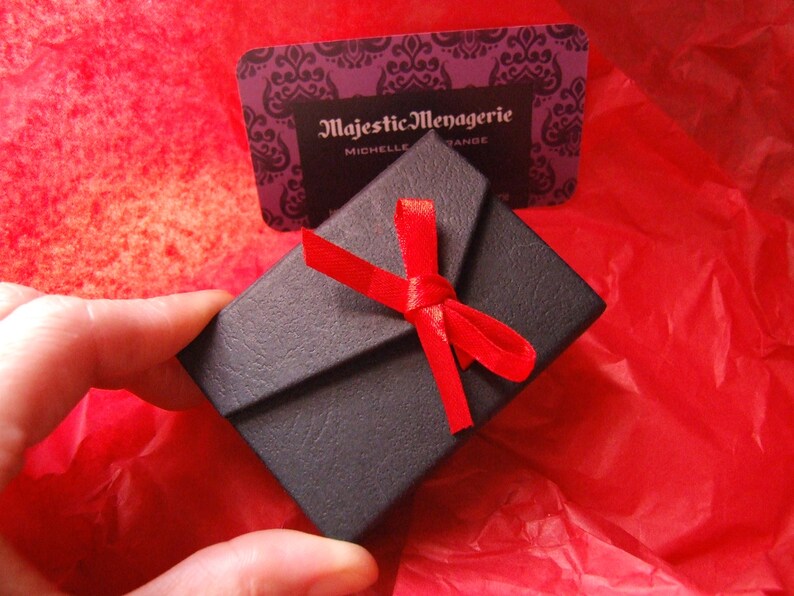 May include: A small black gift box with a red ribbon tied in a bow. The box is sitting on a red tissue paper background. The box has the text "Majestic Menagerie" printed on it.