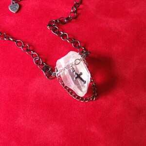 May include: A silver cross charm hangs from a black chain necklace with a clear quartz crystal pendant. The necklace is displayed on a red velvet background.