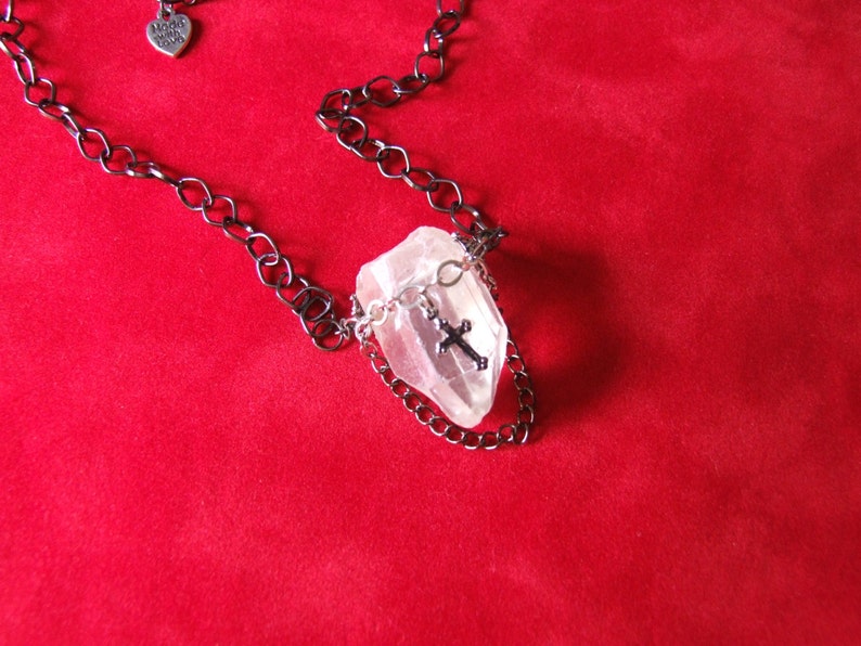 May include: A silver cross charm hangs from a black chain necklace with a clear quartz crystal pendant. The necklace is displayed on a red velvet background.