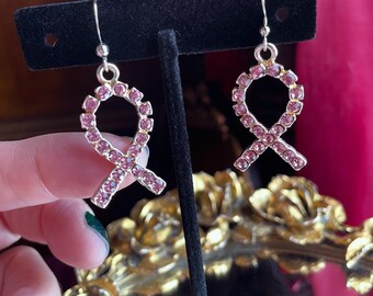 Puede incluir: Un par de pendientes colgantes de tono plateado con un dije en forma de lazo incrustado con pequeños diamantes de imitación de color rosa claro. Los pendientes cuelgan de ganchos plateados simples sobre un soporte negro.