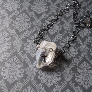 May include: A silver chain necklace with a clear crystal pendant and a small silver cross charm. The necklace is on a gray patterned background.
