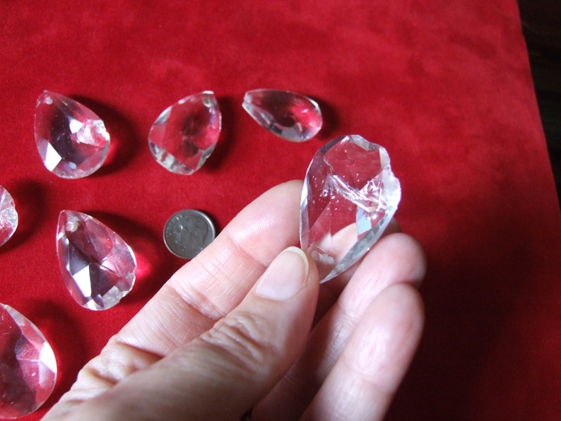 Puede incluir: Una colecci&oacute;n de colgantes de cristal transparente en forma de l&aacute;grima. Los colgantes son facetados y tienen un alto brillo. Se muestran sobre una superficie de terciopelo rojo.
