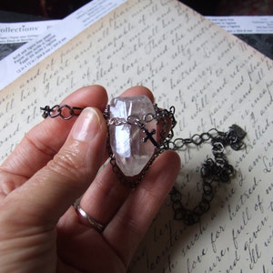 May include: A clear quartz crystal pendant with a silver cross charm hangs from a black chain. The pendant is held in a hand against a background of handwritten text.