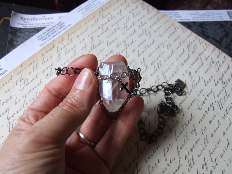May include: A clear quartz crystal pendant with a silver cross charm hangs from a black chain. The pendant is held in a hand against a background of handwritten text.