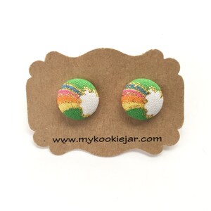 May include: A pair of colorful fabric-covered button earrings. Each earring features a green, white, and rainbow-colored design with gold glitter accents. The earrings are displayed on a brown card with the website address www.mykookiejar.com.