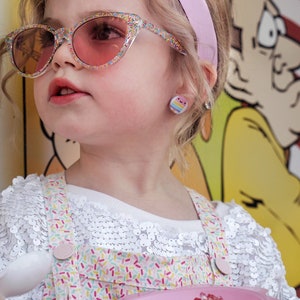 May include: A young girl wearing pink sunglasses and a white shirt with a colorful sprinkle pattern, holding a pink bowl with red shaved ice.