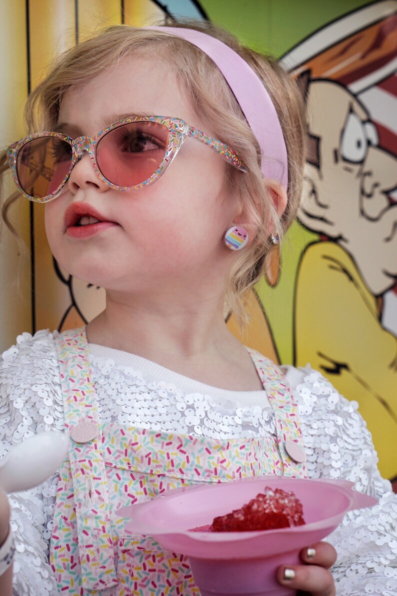 May include: A young girl wearing pink sunglasses and a white shirt with a colorful sprinkle pattern, holding a pink bowl with red shaved ice.