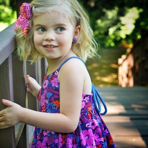 May include: A young girl with blonde hair wearing a purple and pink floral dress with a pink bow in her hair. She is standing on a wooden bridge and looking to the right.