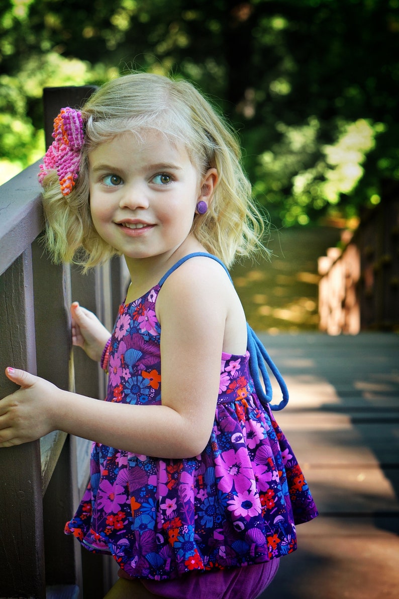 May include: A young girl with blonde hair wearing a purple and pink floral dress with a pink bow in her hair. She is standing on a wooden bridge and looking to the right.