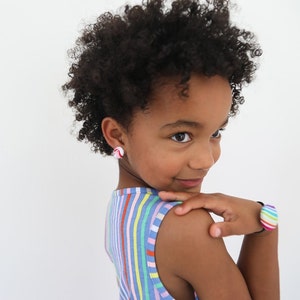 May include: A young girl with curly brown hair wearing a striped sleeveless dress and colorful bracelets. She is wearing a pair of red and white stud earrings.