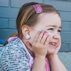 May include: A young girl with blonde hair is wearing a pink and white checkered button earring and a pink glitter hair clip. She is smiling and has her hands on her cheeks.