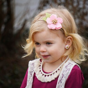 May include: A young girl with blonde hair wearing a pink felt flower headband with green leaves. She is wearing a burgundy shirt with a white lace vest and a white tassel necklace. She is also wearing pink heart-shaped earrings.