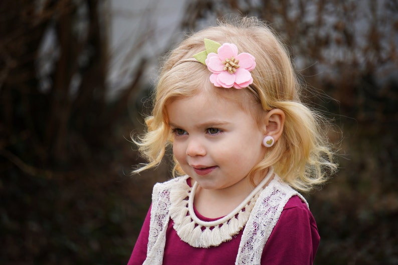 May include: A young girl with blonde hair wearing a pink felt flower headband with green leaves. She is wearing a burgundy shirt with a white lace vest and a white tassel necklace. She is also wearing pink heart-shaped earrings.
