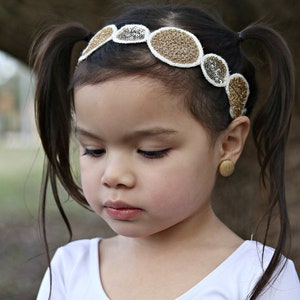 May include: A young girl with brown hair in pigtails wears a white headband with gold and silver embellishments. The headband is made of fabric and has a circular design.