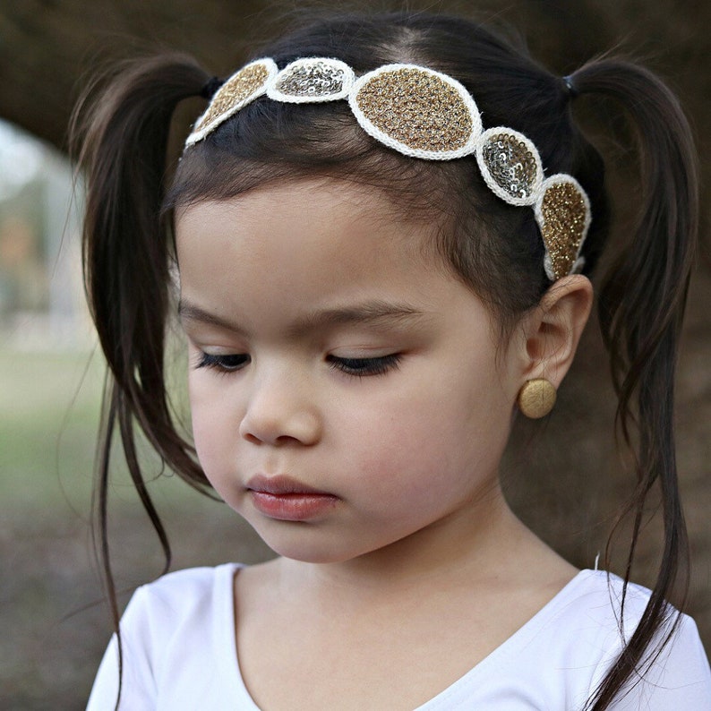 May include: A young girl with brown hair in pigtails wears a white headband with gold and silver embellishments. The headband is made of fabric and has a circular design.