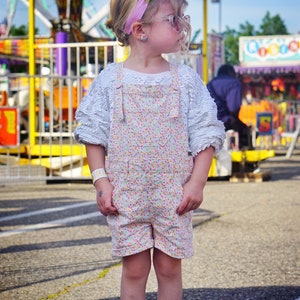 May include: A young girl wearing a white, lacy, long-sleeved shirt with a floral print overall dress and pink sunglasses. She is standing in front of a yellow and purple carnival ride.
