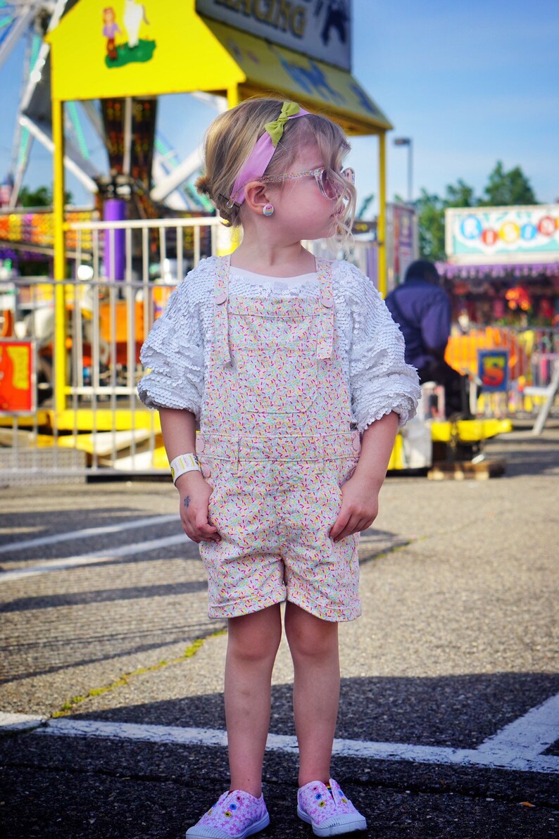 May include: A young girl wearing a white, lacy, long-sleeved shirt with a floral print overall dress and pink sunglasses. She is standing in front of a yellow and purple carnival ride.