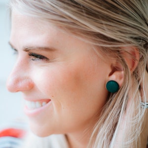 May include: A close-up of a woman's ear with a pair of dark green round stud earrings. The earrings are made of a smooth, matte material.