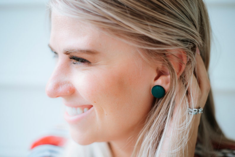 May include: A close-up of a woman's ear with a pair of dark green round stud earrings. The earrings are made of a smooth, matte material.