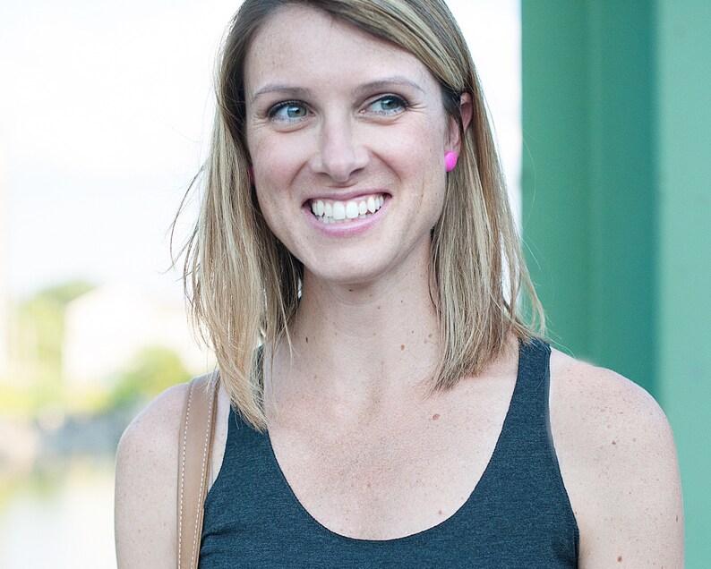 May include: A woman with blonde hair smiling and wearing a black tank top and pink earrings.