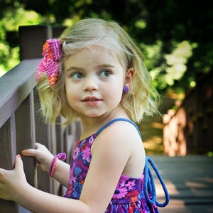 May include: A young girl with blonde hair wearing a purple and pink floral dress with a blue strap. She is wearing a pink flower in her hair and purple earrings. She is standing next to a wooden railing.